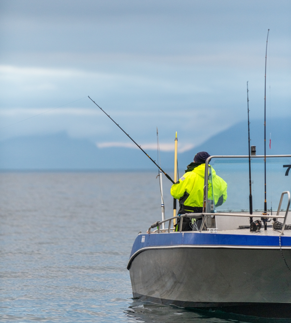 Fisker i gul jakke står på båt og fisker. Foto.