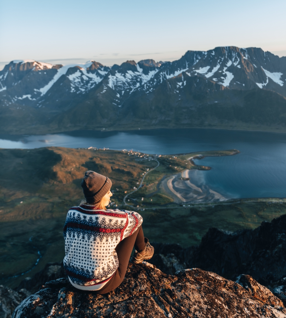 Jente med lue sitter på fjell og ser utover havet og Nuvsvåg. Foto.