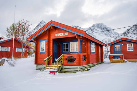 Tre røde hytter med blå dører og karmer. Snødekt landskap. Foto.