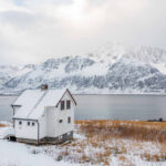 Hvitt hus i vinterlandskap med hav og fjell i bakgrunnen. Foto.