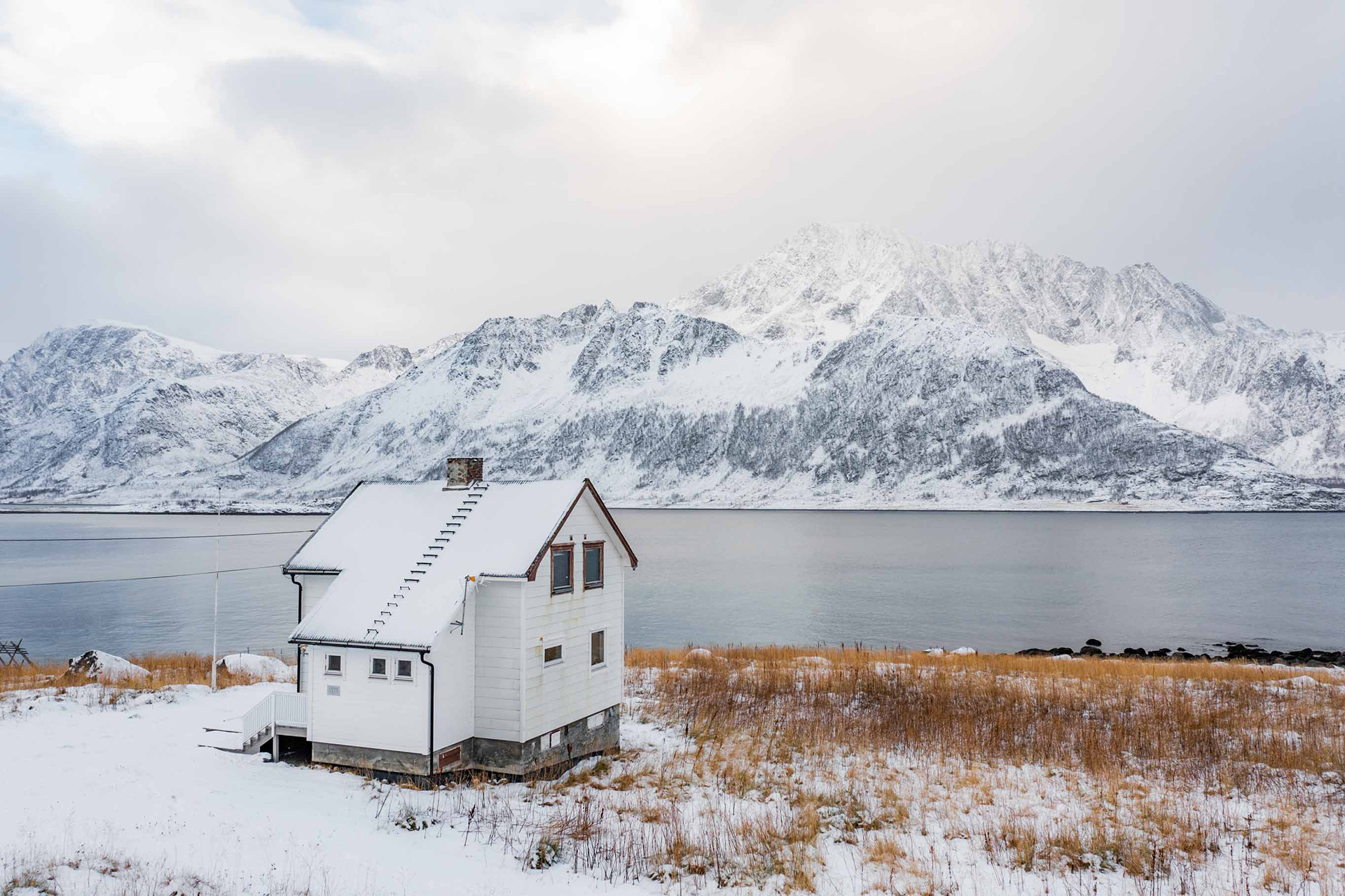 Hvitt hus i vinterlandskap med hav og fjell i bakgrunnen. Foto.