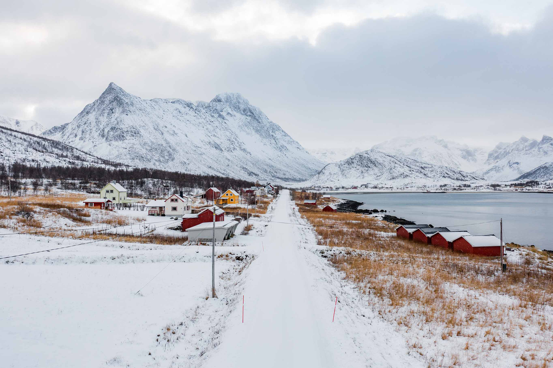 Vei og hus langs veien. Fjell med snø i bakgrunnen. Foto.