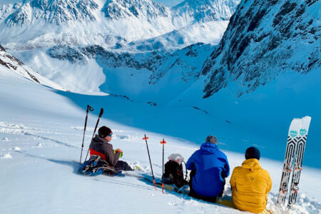 Randonee topptur med familie sett bakfra, spiser lunsj på snødekt fjell.