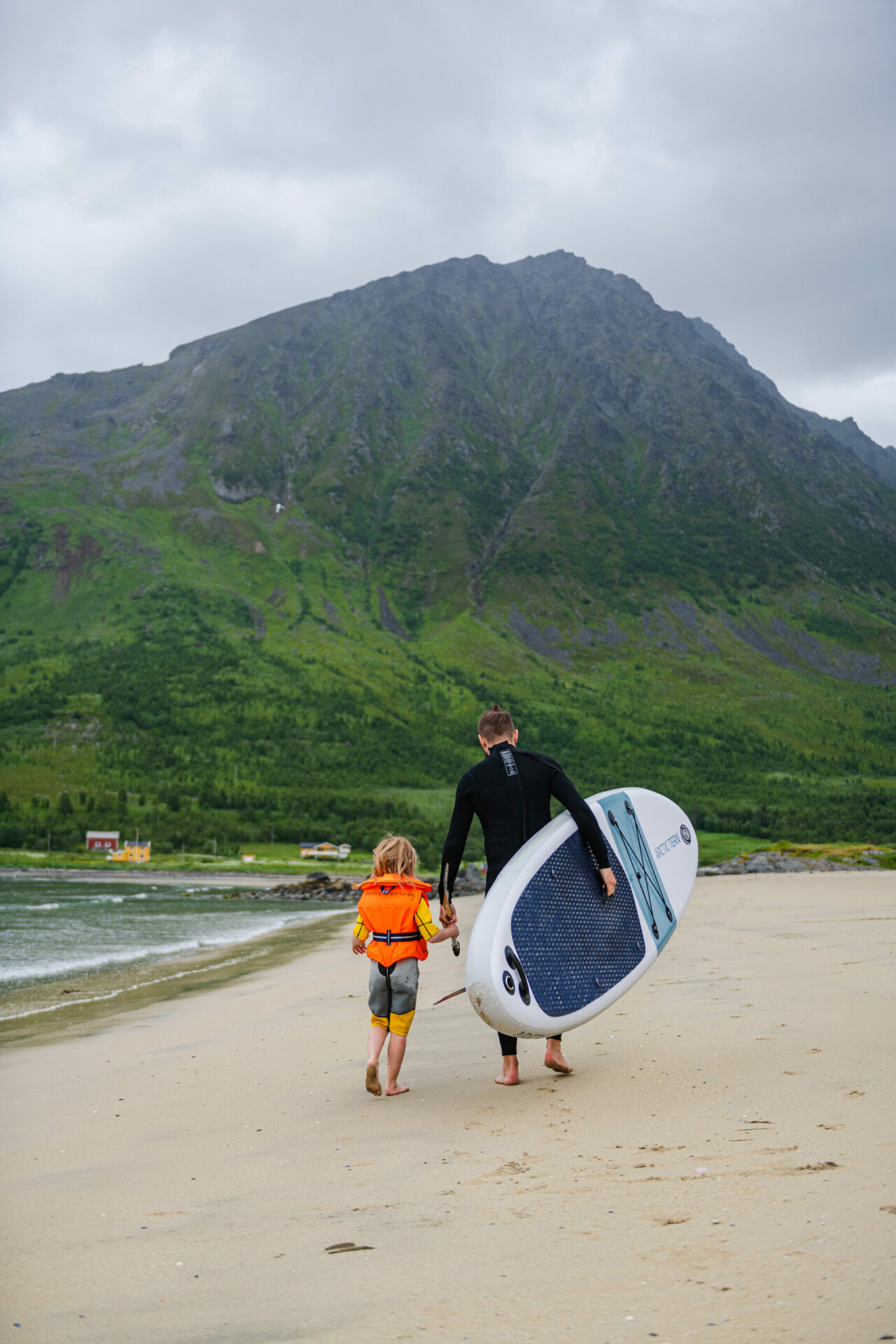 Mann og barn går lamgs strand med sup-brett og årer under armene. Foto.